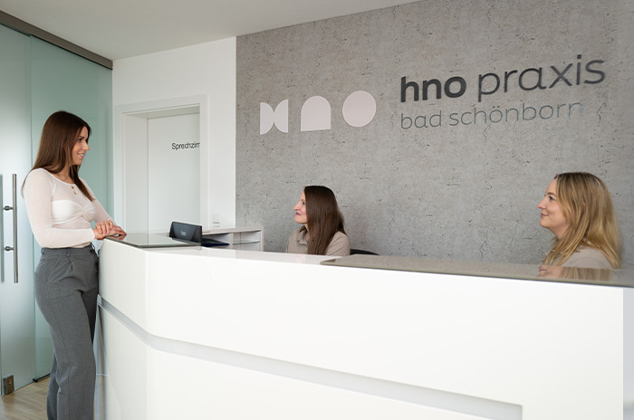 Zwei am Empfangstresen sitzenden Mitarbeiterinnen im Gespräch mit einer am Tresen stehenden Patientin